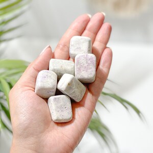 May include: A hand holding a collection of small, white, square-shaped stones with purple and pink veins. The stones are smooth and polished.