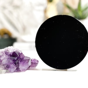 May include: A black obsidian mirror and a cluster of amethyst crystals on a white surface.
