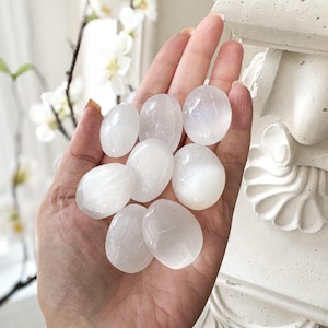 May include: A hand holds a collection of smooth, oval-shaped selenite stones. The stones are translucent white, with a subtle sheen. The background includes white flowers and a decorative architectural element.