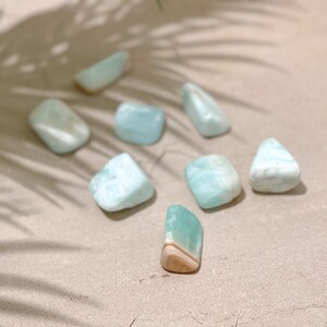 May include: A collection of seven light blue and white aquamarine tumbled stones on a beige surface. The stones are smooth and polished, with a variety of shapes and sizes.