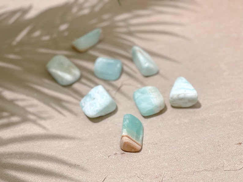 May include: A collection of seven light blue and white aquamarine tumbled stones on a beige surface. The stones are smooth and polished, with a variety of shapes and sizes.