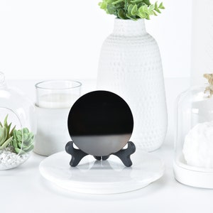 May include: A black circular mirror on a black stand, with a white base. The mirror is on a white surface with a glass cloche, a glass globe with succulents, and a white vase with green plants in the background.