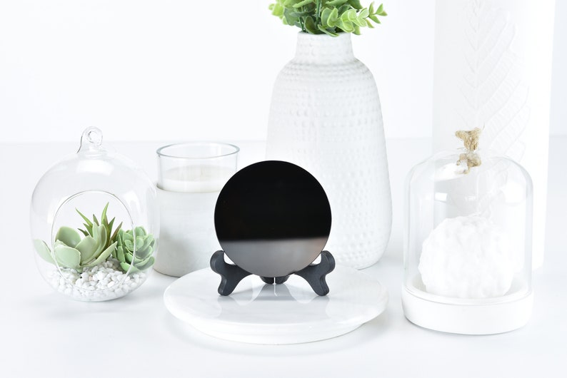 May include: A black circular mirror on a black stand, with a white base. The mirror is on a white surface with a glass cloche, a glass globe with succulents, and a white vase with green plants in the background.