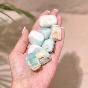 May include: A collection of tumbled blue and green aquamarine stones with white and brown inclusions. The stones are smooth and polished, and they are held in a person's hand.