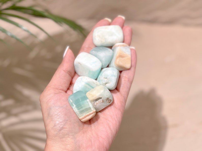 May include: A collection of tumbled blue and green aquamarine stones with white and brown inclusions. The stones are smooth and polished, and they are held in a person's hand.