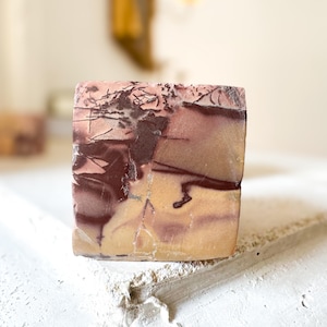 May include: A square-shaped stone with a unique pattern of colors. The stone features a blend of light pink, beige, and dark brown hues, creating a marbled effect. The stone is likely a decorative item or a piece of natural art.