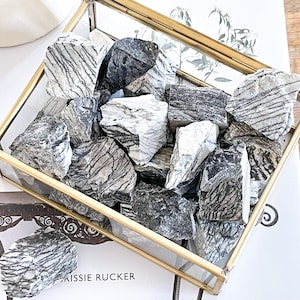 May include: A glass box filled with black and white striped stones. The box is made of gold metal and has a glass lid. The stones are arranged in a random pattern.