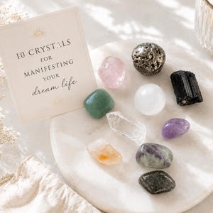 May include: A collection of ten assorted polished and raw crystals arranged on a round white stone tray, accompanied by a square card with the text 10 CRYSTALS FOR MANIFESTING YOUR dream life, alongside a small beige fabric drawstring pouch.