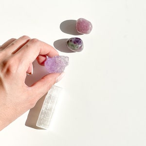 May include: A collection of purple and green crystals, including amethyst, selenite, and fluorite, on a white background.
