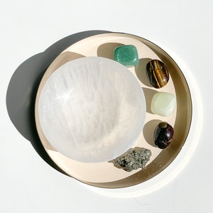 May include: A white selenite bowl with a variety of crystals including green aventurine, tiger's eye, green jade, red jasper, and pyrite on a gold tray.