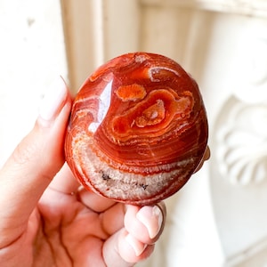 May include: A polished, oval agate stone in red and orange hues. The stone displays concentric bands and patterns, with a white and grey crystalline centre. The gemstone is held in a hand.