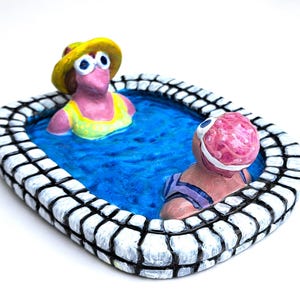 May include: A whimsical ceramic sculpture of two figures in a blue pool. One figure wears a yellow hat and swimsuit, the other a pink swim cap and purple swimsuit. The pool has a white and black brick border.