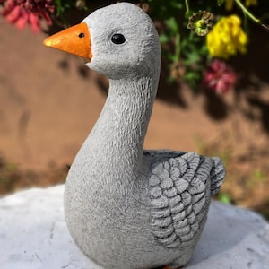 May include: A gray goose figurine with an orange beak and feet. The goose has a textured surface and is positioned in front of a blurred background of flowers. This decorative item is suitable for garden or home decor.