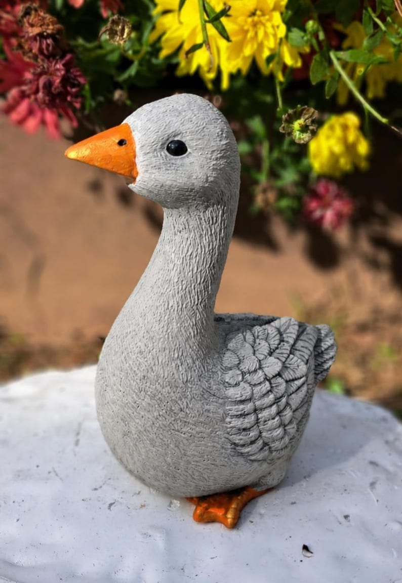 May include: A gray goose figurine with an orange beak and feet. The goose has a textured surface and is positioned in front of a blurred background of flowers. This decorative item is suitable for garden or home decor.