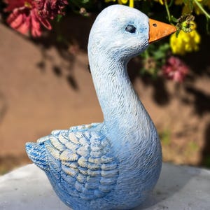 May include: A light blue goose figurine with an orange beak and feet. The goose has a textured surface with white and gold accents on its wings. The figurine is a decorative item, possibly for a garden or home decor.