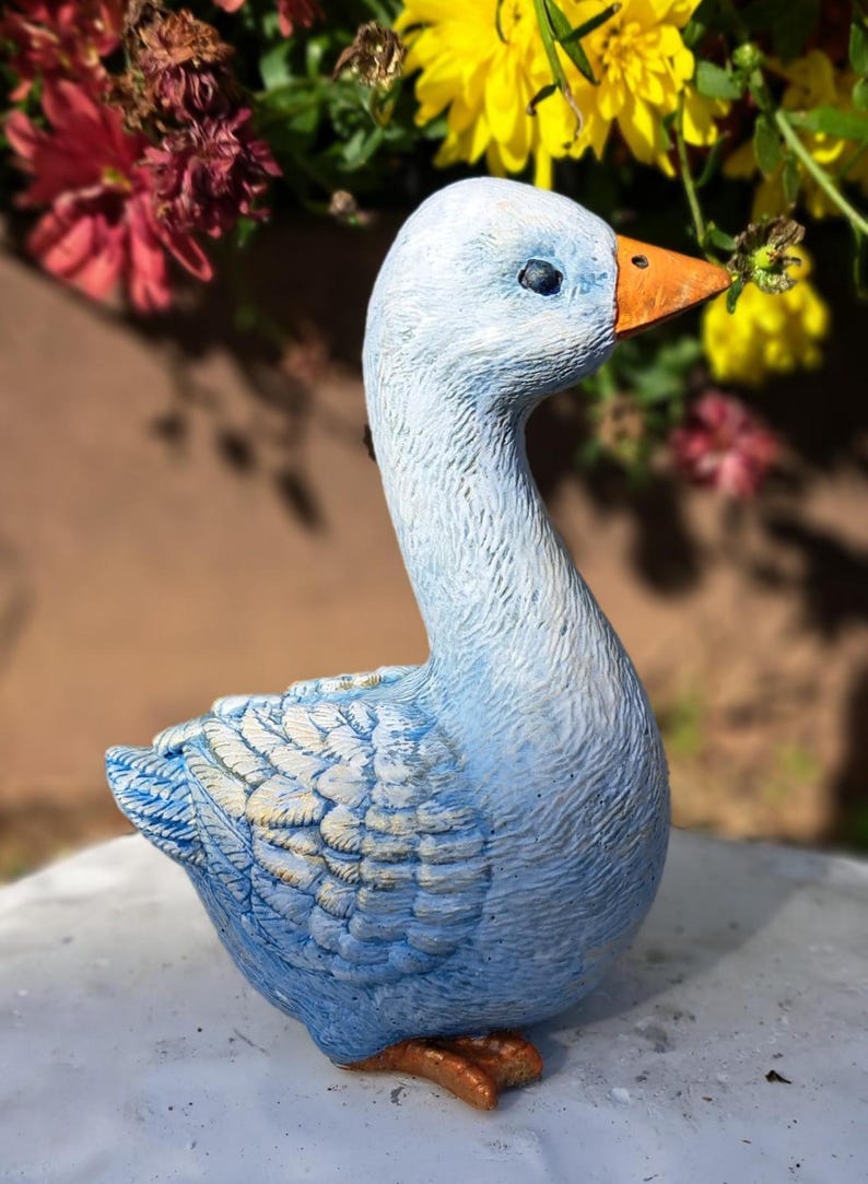 May include: A light blue goose figurine with an orange beak and feet. The goose has a textured surface with white and gold accents on its wings. The figurine is a decorative item, possibly for a garden or home decor.