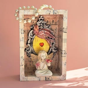May include: A decorative shadow box featuring a bird, angel, and heart design. The artwork includes a bird with red and white wings, a yellow heart, and a small angel holding flowers. A pearl necklace is draped over the top.