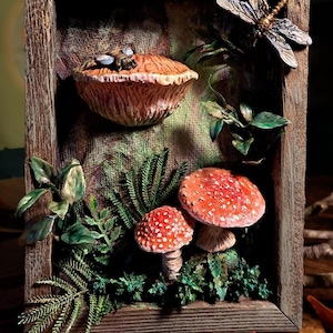 May include: A framed woodland scene featuring detailed mushroom sculptures with red caps and white spots, surrounded by green foliage and a dragonfly. The frame is made of wood, creating a natural aesthetic.
