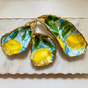 May include: Three gold-painted oyster shells with a lemon and leaf design. The shells are on a white marble surface.
