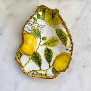 May include: A white ceramic dish shaped like an oyster shell with a gold rim. The dish is decorated with a lemon print featuring yellow lemons and green leaves.