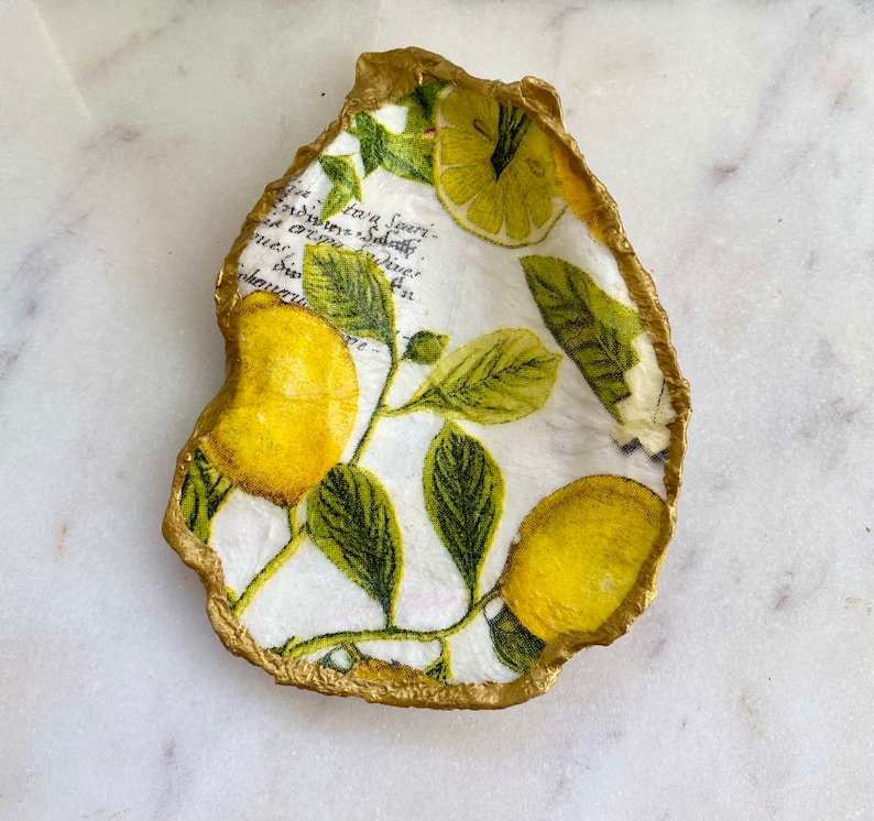 May include: A white ceramic dish shaped like an oyster shell with a gold rim. The dish is decorated with a lemon print featuring yellow lemons and green leaves.