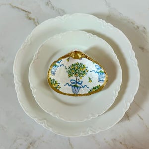 May include: A decorative seashell with a gold-painted rim and a hand-painted design featuring a lemon tree in a blue pot, set on two white plates. The shell's interior has a white background with blue and gold accents.