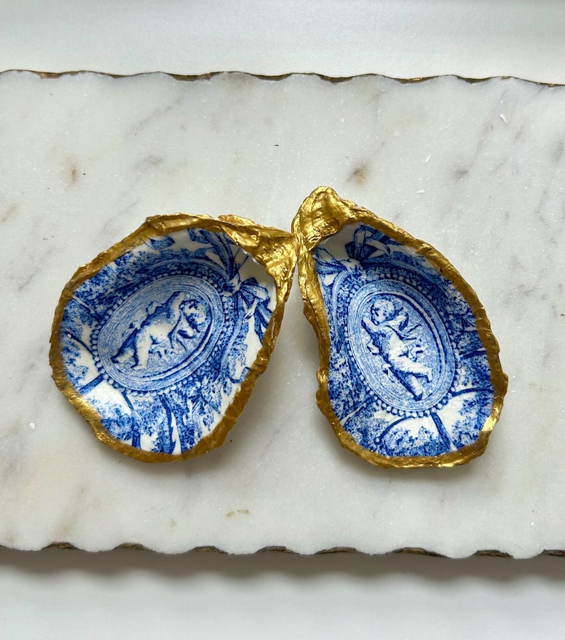 Pu&ograve; includere: Due conchiglie di ostriche in porcellana blu e bianca con finiture dorate. Ogni conchiglia presenta un design di cherubino.