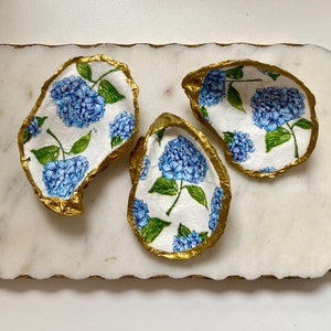 May include: Three white oyster shells with gold trim, each painted with blue hydrangeas and green leaves.