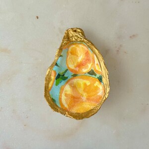 May include: A gold-painted oyster shell with a blue and white pattern of oranges. The shell is a decorative dish or trinket.