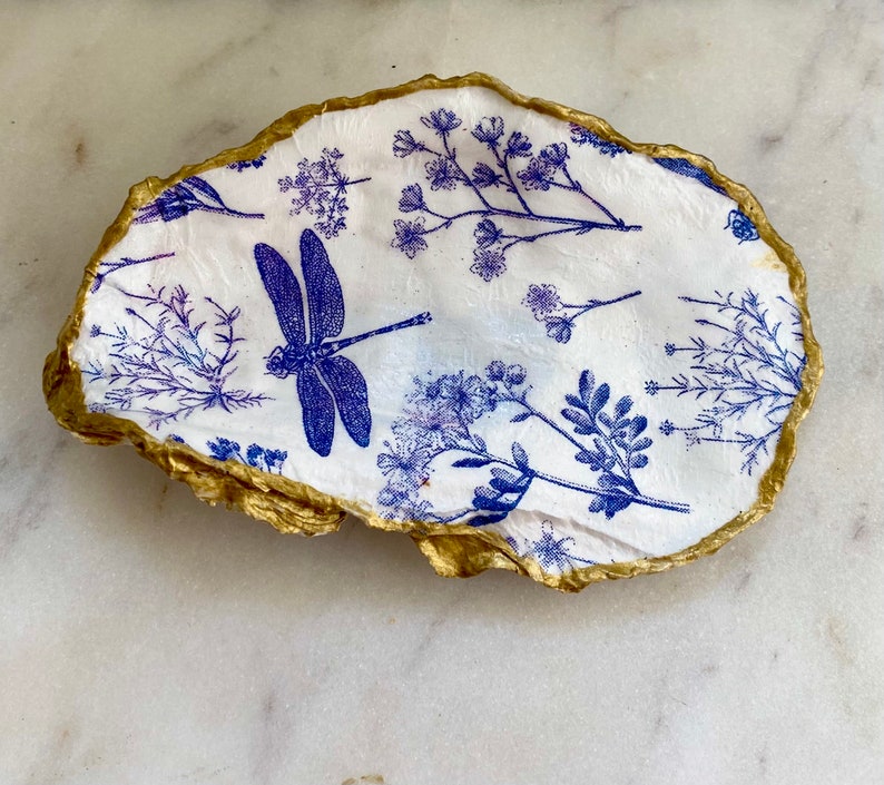 May include: A white oyster shell dish with a gold rim. The dish is decorated with a blue dragonfly and blue floral patterns.