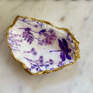 May include: A white oyster shell dish with a gold rim. The dish is decorated with a purple dragonfly and purple floral designs.