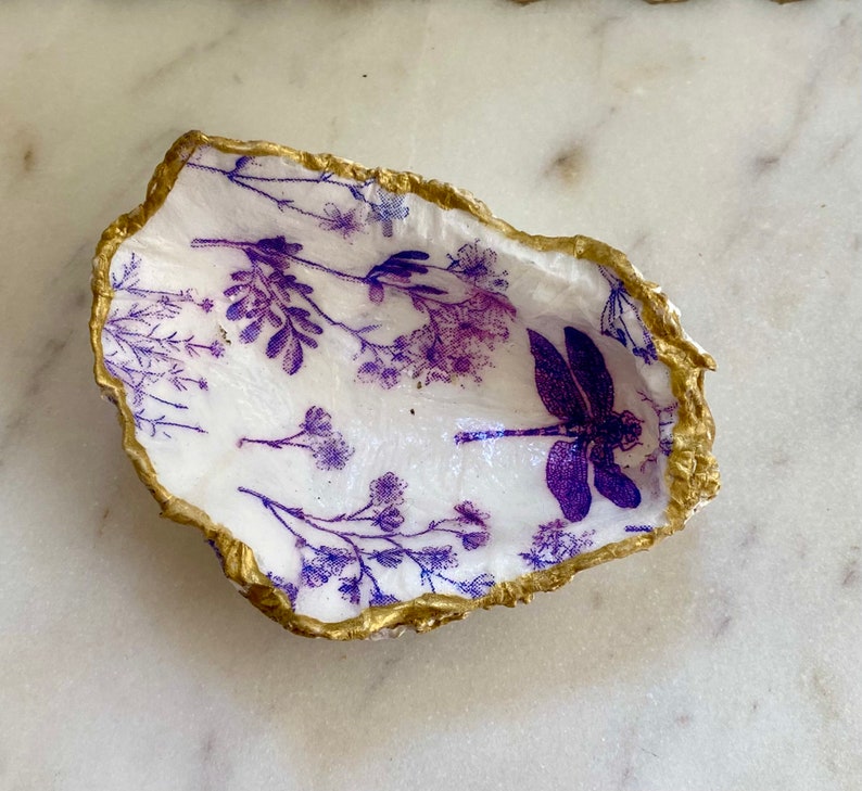 May include: A white oyster shell dish with a gold rim. The dish is decorated with a purple dragonfly and purple floral designs.