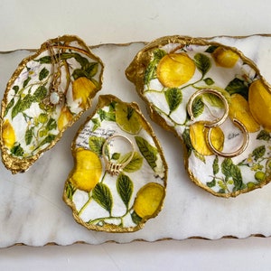 May include: Three white ceramic dishes shaped like seashells with gold trim. Each dish features a floral design with green leaves and yellow flowers. The dishes are decorated with gold accents and are perfect for holding jewelry or other small items.
