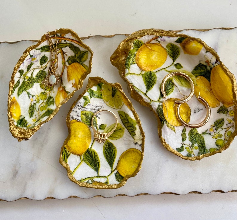 May include: Three white ceramic dishes shaped like seashells with gold trim. Each dish features a floral design with green leaves and yellow flowers. The dishes are decorated with gold accents and are perfect for holding jewelry or other small items.