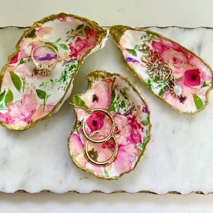 May include: Three decorative oyster shell dishes with a floral pattern in shades of pink, white, and green. The shells are trimmed with gold paint and are used to hold jewelry.