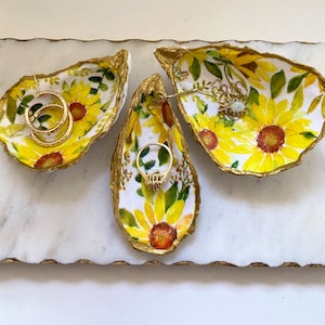 May include: Three white ceramic oyster shell dishes with gold trim and a sunflower pattern. The dishes are filled with jewelry, including a pair of gold hoop earrings and a gold ring.