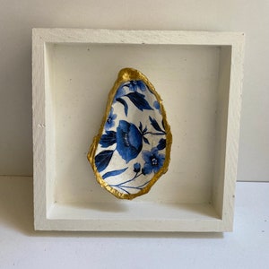 May include: A white oyster shell with a blue and white floral pattern is framed in a white wooden frame. The shell is painted with gold around the edges.