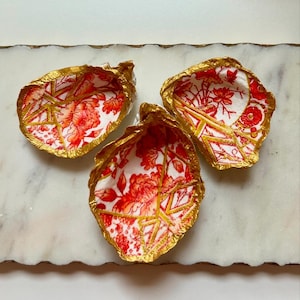 May include: Three decorative seashells with a red and gold floral pattern. The shells are painted with a gold trim and have a white background.