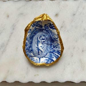 Pu&ograve; includere: Un guscio di ostrica in porcellana blu e bianca con un bordo dorato. Il guscio presenta un design decorativo con un ovale centrale che contiene una figura in un motivo floreale blu e bianco.