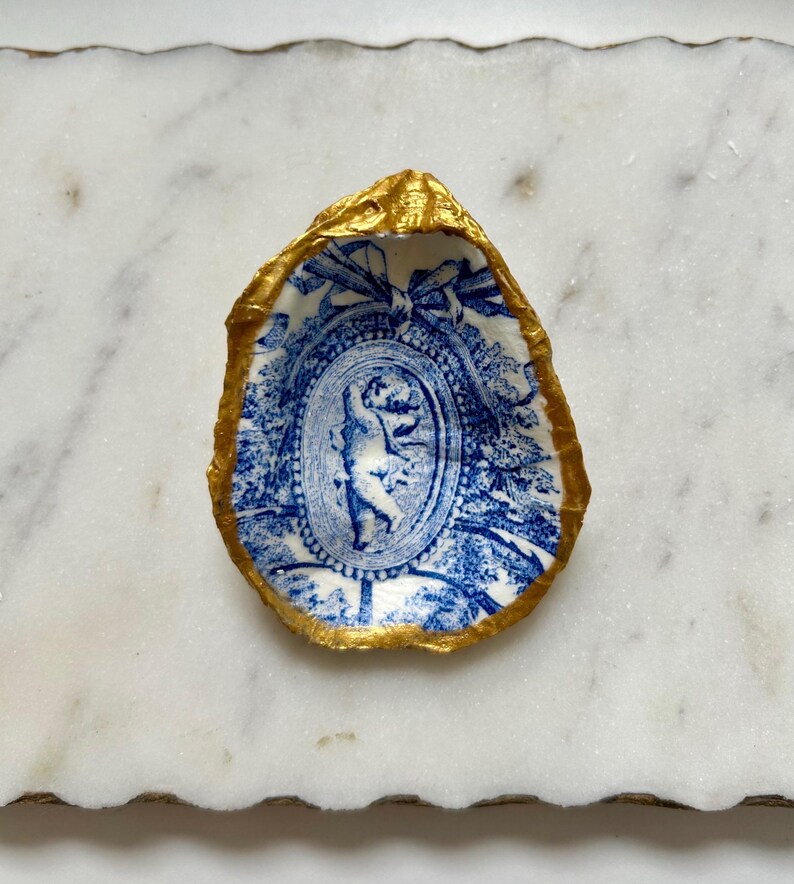 Pu&ograve; includere: Un guscio di ostrica in porcellana blu e bianca con un bordo dorato. Il guscio presenta un design decorativo con un ovale centrale che contiene una figura in un motivo floreale blu e bianco.