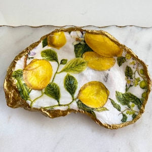 May include: A white ceramic dish with a gold rim, featuring a lemon and leaf design. The dish has a textured surface and is shaped like an oyster shell.