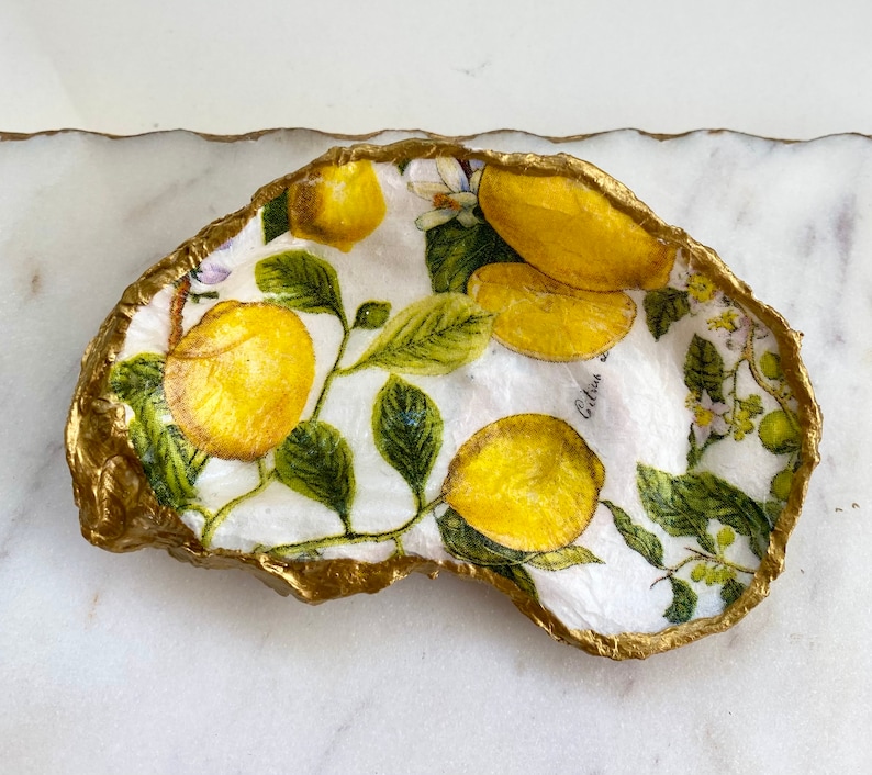 May include: A white ceramic dish with a gold rim, featuring a lemon and leaf design. The dish has a textured surface and is shaped like an oyster shell.