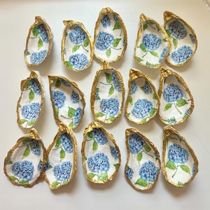 May include: A collection of oyster shells with a gold rim and a floral design featuring blue hydrangeas on a white background. The shells are arranged on a white surface.