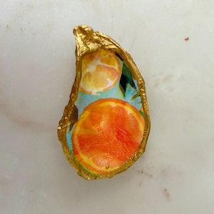 May include: A gold-painted oyster shell with a blue and green background and a design of orange slices.