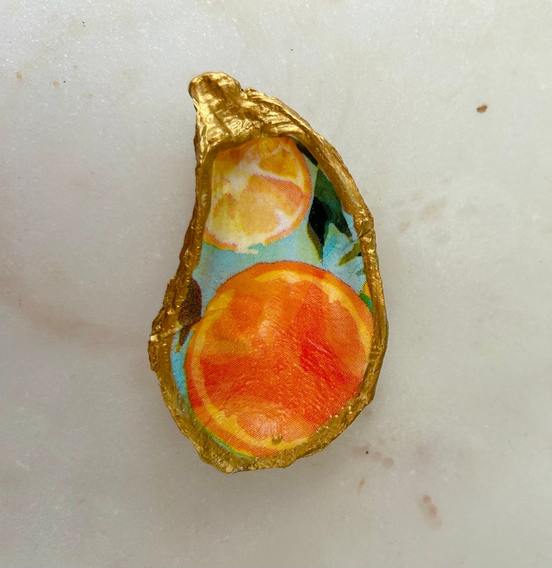 May include: A gold-painted oyster shell with a blue and green background and a design of orange slices.