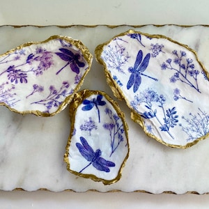 May include: Three decorative oyster shells with a gold rim. Each shell is decorated with a white background and a blue dragonfly and floral pattern.