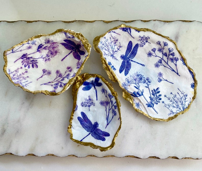 May include: Three decorative oyster shells with a gold rim. Each shell is decorated with a white background and a blue dragonfly and floral pattern.