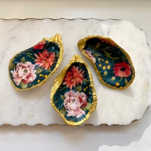 May include: Three decorative oyster shells with gold edges and floral designs. The interiors feature a dark teal background with pink and red flowers and green leaves. The shells are arranged on a white marble surface.