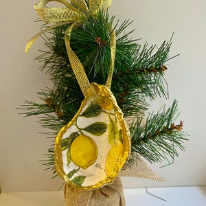 May include: A gold-painted oyster shell ornament with a lemon print and the word "Citrus" on it. The ornament is hanging from a gold ribbon on a small artificial Christmas tree.