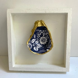 May include: A white wooden frame with a gold-painted oyster shell inside. The oyster shell is decorated with a blue and white floral pattern.